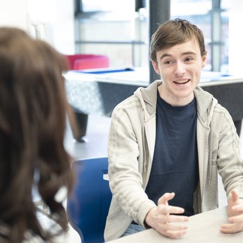 A student speaking to a teacher, sat at a table.