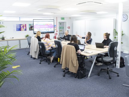 A group of people sit around a cream office table with a presentation screen