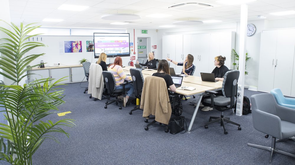 A group of people sit around a cream office table with a presentation screen