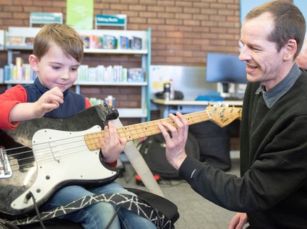 A boy strums a electric guitar with the help of a teacher in the library