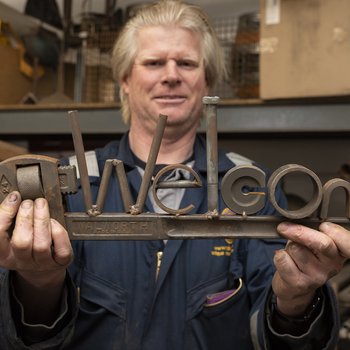 White man with blonde hair holding up welded iron letters saying 'welcome'