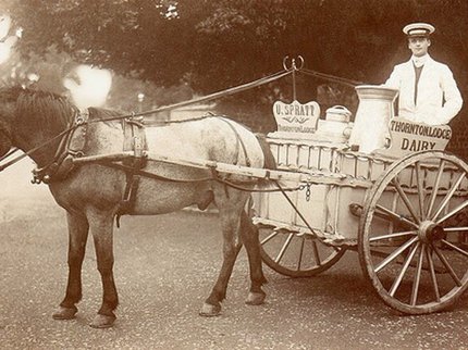 A man standing on a horse-drawn dairy cart beside a young child