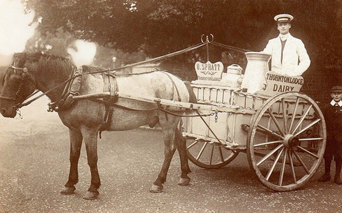 A man standing on a horse-drawn dairy cart beside a young child