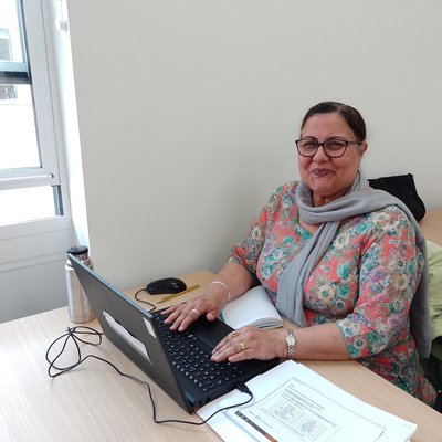 A woman with glasses, a flowery top and grey scarf smiles while she uses a laptop