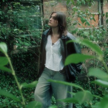 A young woman stood against a leafy background wearing jeans and a leather-look jacket