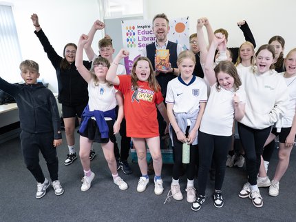 A group of children stand with author Alastair Chisholm and smile with their hands raised in the air