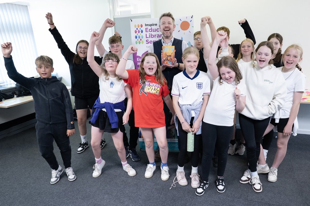 A group of children stand with author Alastair Chisholm and smile with their hands raised in the air