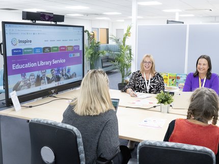 A group of people sit at a large desk and talk next to a large digital screen