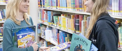 Two women holding books stand and talk in front of book shelves