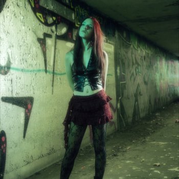 A young woman with long dark hair stood against a wall of graffiti, wearing black tights and boots with a red skirt.