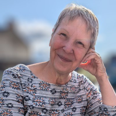 An older white woman with short grey hair is smiling at the camera whilst leaning on her hand.