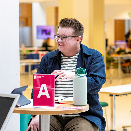 A young man sat at a desk smiling looking to the side.