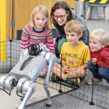 A woman and three young children smiling whilst looking at a robot dog.