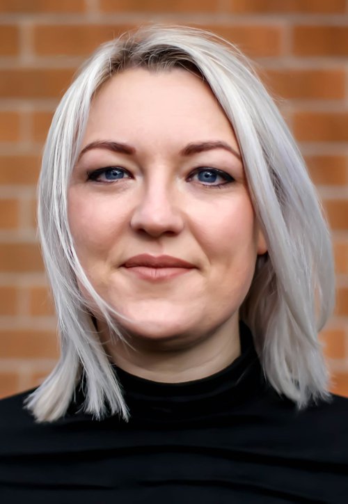 A headshot of a young woman with short light blonde hair and blue eyes, wearing a black top.