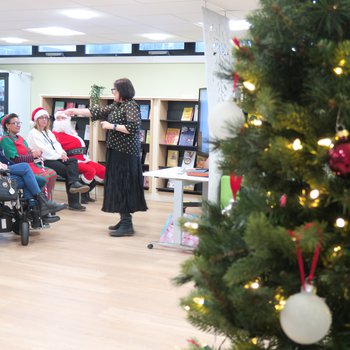 Santa and his elf sat watching someone sing, beside a Christmas tree
