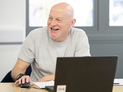 A middle aged man wearing a light grey t-shirt sits at a computer, holding a mouse whilst smiling. The man is white with a bald head, moustache and goatee beard.