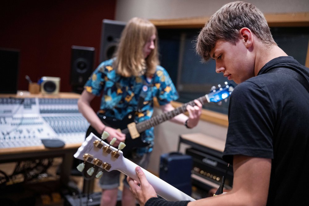 A band playing on stage. 2 people are at the front, one is playing guitar and one is singing into a microphone. There is a person in the background playing drums.