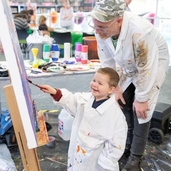a young boy painting at an easel with an an older male artist standing behind him