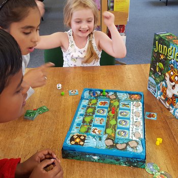 One boy and two girls, aged between 5 and 8, are playing a board game called Jungle Party. One girl is raising both hands in celebration and smiling, the other two children are looking at the board, their facial expressions engaged and interested.