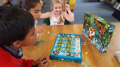 One boy and two girls, aged between 5 and 8, are playing a board game called Jungle Party. One girl is raising both hands in celebration and smiling, the other two children are looking at the board, their facial expressions engaged and interested.