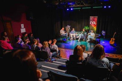 A black and white image of a guitarist, a woman leaning against a piano and a man playing the piano and smiling.