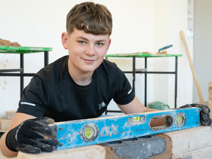 A construction student smiles at the camera whilst checking that a brick wall is level