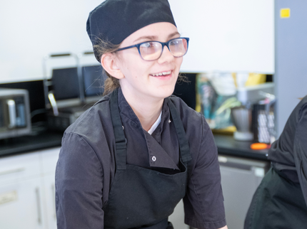 A young student is wearing a sous-chef uniform and cooking in a kitchen. They smile as they work.
