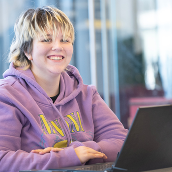 A younger student with short blonde hair smiles from behind a laptop