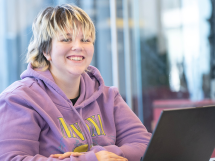 A student in a light purple hoodie is using their laptop and smiling at the camera