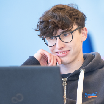 A young student with dark curly short hair and glasses is using a laptop