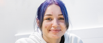 A young Inspire College student with purple hair in a ponytail is smiling at the camera