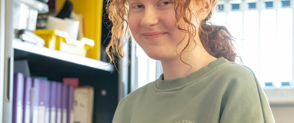 A teenage girl with curly ginger hair in a low pony tail and wearing a sage green jumper smiling.