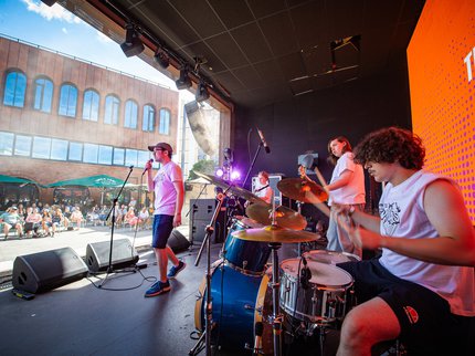 A young band performing on stage, looking out at the audience. There is a drummer, singer and 2 guitarists within the image. The sun is shining and there are around 50 people pictured in the audience at Binks Yard in Nottingham.