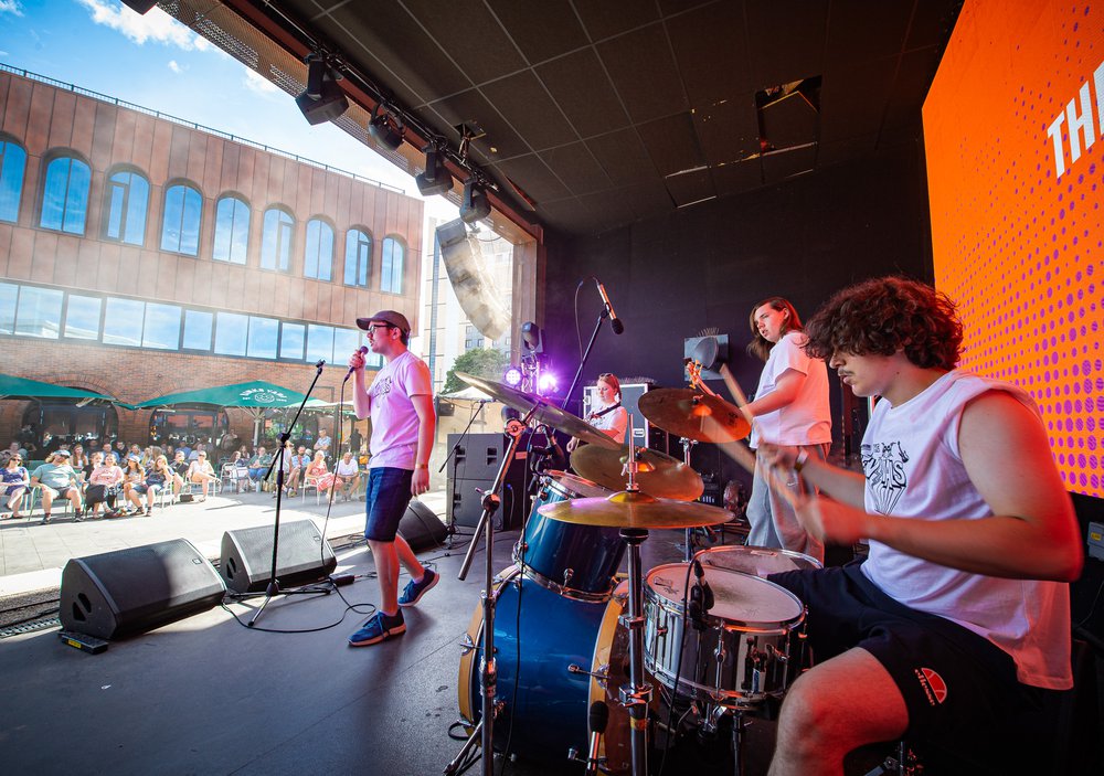 A young band playing at an event. There is a drummer, singer and 2 guitarists in the shot. They are playing to an audience of around 50 people at Binks Yard in Nottingham.