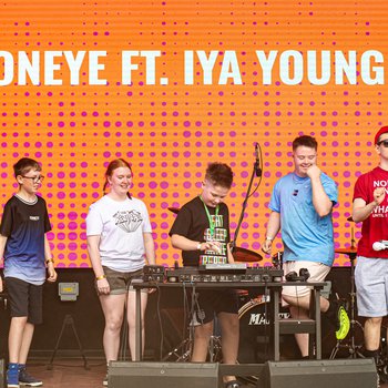 A group of young people on a stage with an orange background, the boy in the middle is DJing