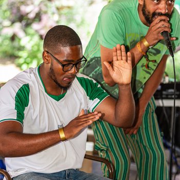 A man drumming while another man sings into a microphone. Both are wearing green