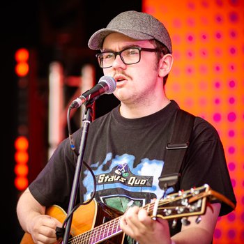 A boy wearing a cap singing and playing guitar on stage