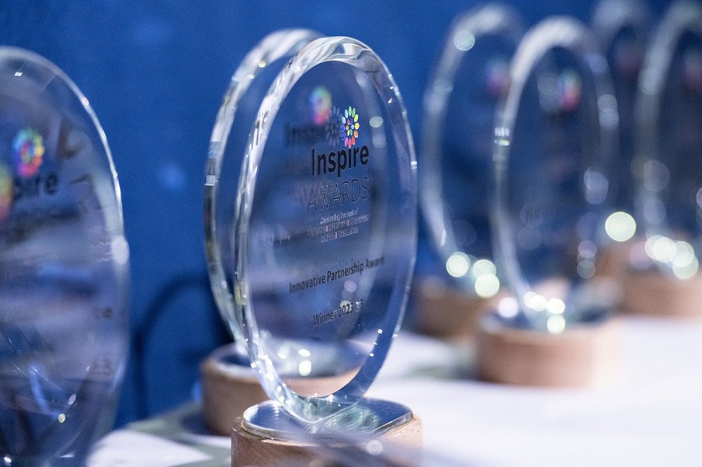 Inspire Awards glass trophies sit on a table with a blue backdrop ready for their winners.