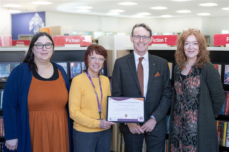 Group of 4 smiling people, 3 female and 1 male in a library setting receiving a certificate