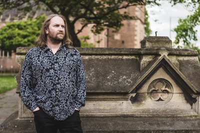 Historian James Wright - a white male with long, light brown hair and a short beard - stands in front of a sandstone tomb