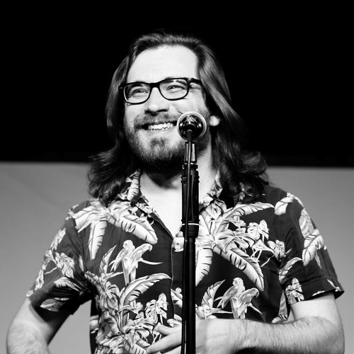 A black and white headshot image of a person wearing glasses stood in front of a microphone.