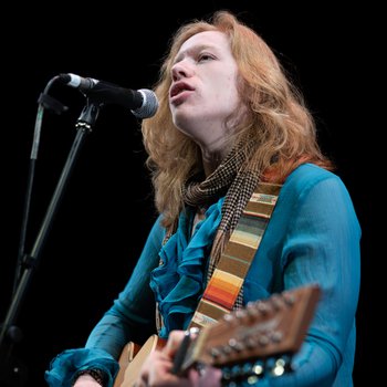 A man with long red hair playing the acoustic guitar and singing. He is wearing a blue shirt.