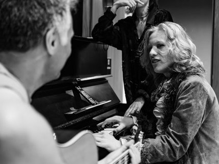 A black and white image of two musicians, one sits at a piano and the other strums a guitar