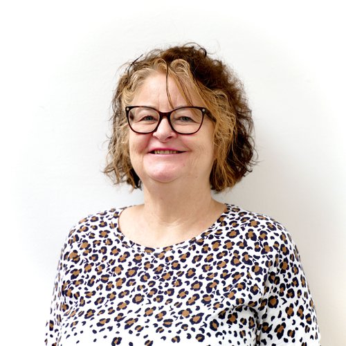 Female with short brown and blonde curly hair, wearing black framed glasses, smiling to camera. Wearing a white, black and brown leopard print top, standing against a white background,