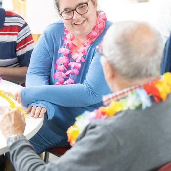 A college student wearing flowers around her neck, talking to an elderly resident.