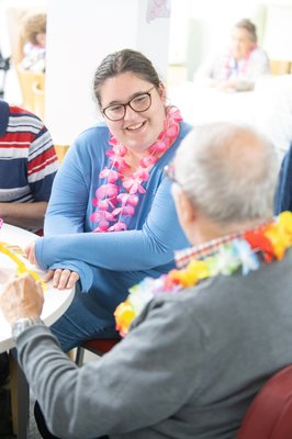 A college student wearing flowers around her neck, talking to an elderly resident.