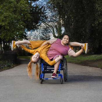 A curvy brown skinned filipino female wheelchair dancer, with an above right knee amputation sat in a manual wheelchair, she leans to the left, her left hands holds the ankle of another woman whose leg rest on her shoulders. She wears a plum coloured dress. Her right arm is clasped around the waist of a white woman with ginger hair who hangs upside down with her legs in a split. The woman with ginger hair holds her left ankle and has her left hand on the first woman’s left knee, she wears an ochre jumpsuit.