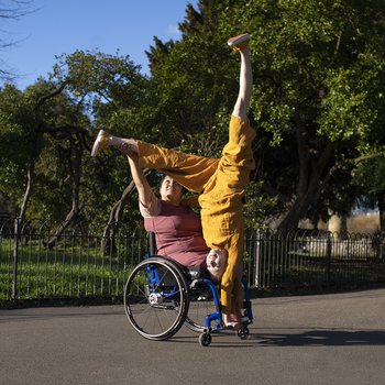 A curvy brown skinned filipino female wheelchair dancer, with an above right knee amputation sat in a manual wheelchair. She wears a plum coloured dress, her right hand holding up the left leg of a white woman with ginger hair who does a one-armed handstand on the footrest of the wheelchair. The woman with ginger hair wears an ochre jumpsuit with her feet flexed, one leg reaching up and the other held by the wheelchair dancer. They look out towards the sunlight smiling. They are on a grey sunlit asphalt pat