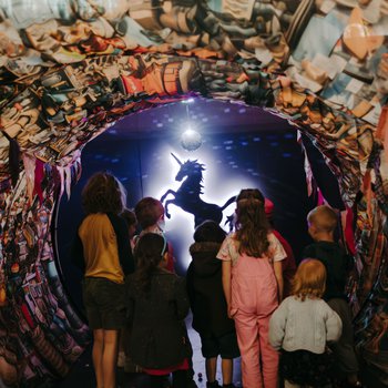 A group of children are gathered together, looking through a tunnel made of colourful, textured fabric. They are looking at a silhouette of a unicorn in front of a bright light.