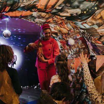 A performer in a red jumpsuit and hat is holding a puppet, interacting with a group of children. The scene is illuminated by a disco ball hanging from the ceiling of a decorated fabric tunnel.
